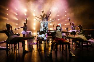 Balettiasuisia tanssijoita Billy Elliot -musikaalin kohtauksessa, osa istuu ja osa seisoo. Edessä istuu poika nyrkkeilyasussa.