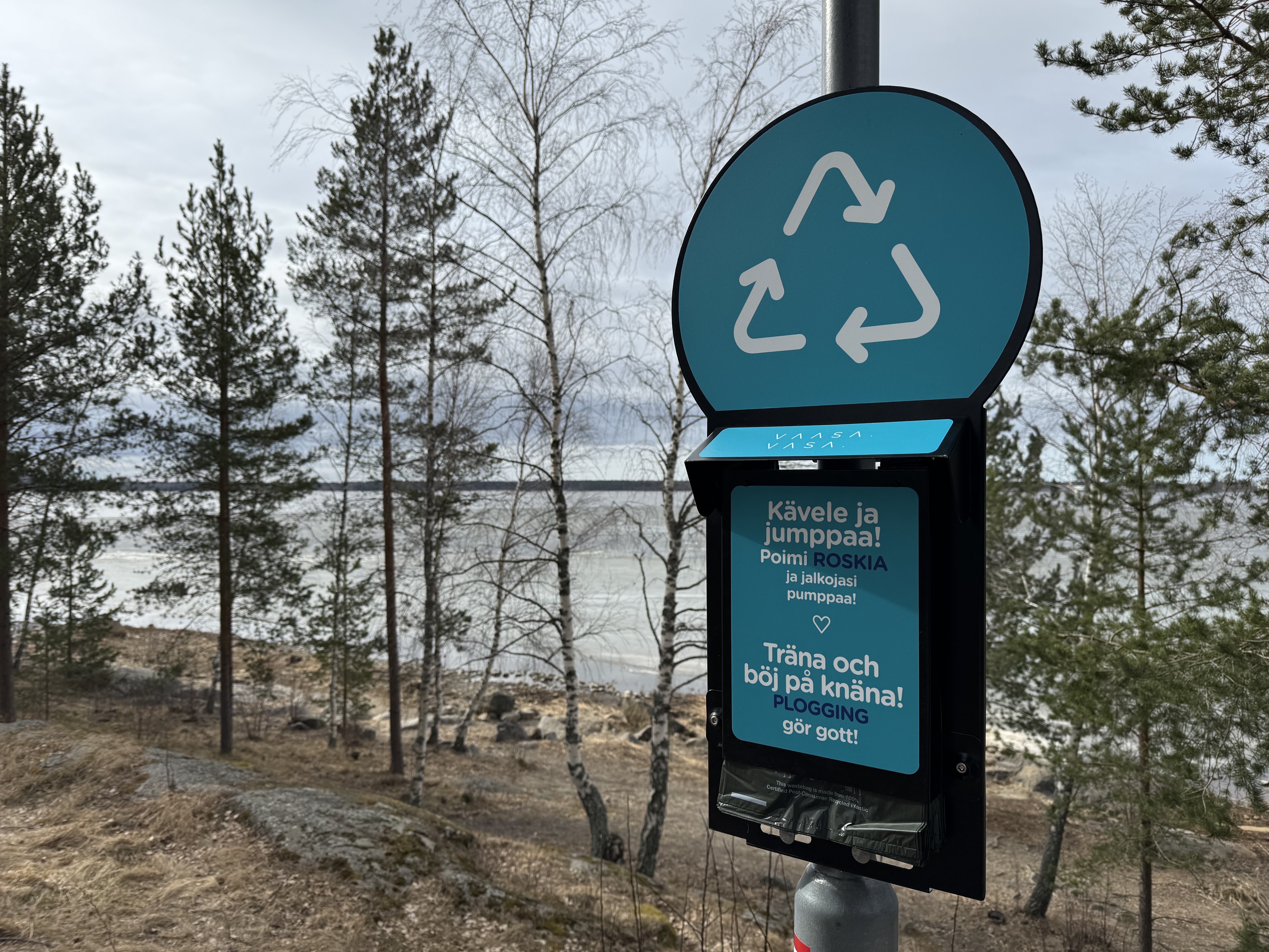 Sinisin kyltein varustettu roskapussiteline meren rannalla puistossa.