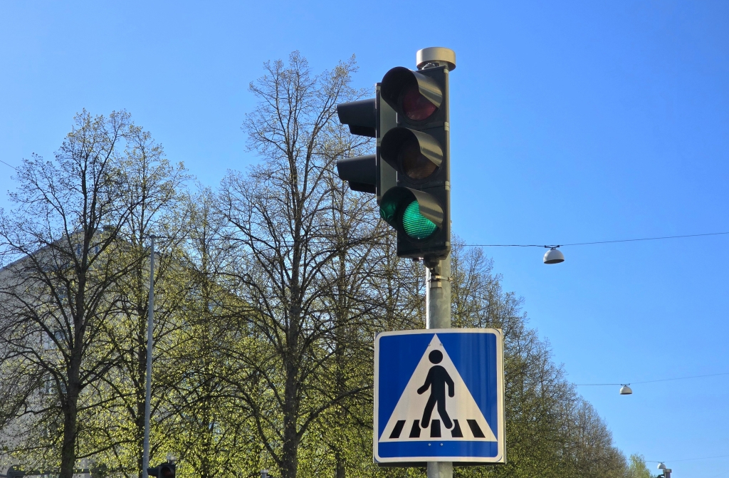 Liikennevalo näyttää vihreää valoa, ja sen alla on suojatien merkki. Taustalla on lehtipuita.
