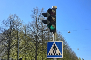 Liikennevalo näyttää vihreää valoa, ja sen alla on suojatien merkki. Taustalla on lehtipuita.