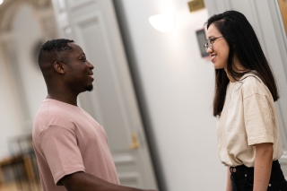 Two people are standing indoors facing each other and smiling at one another.