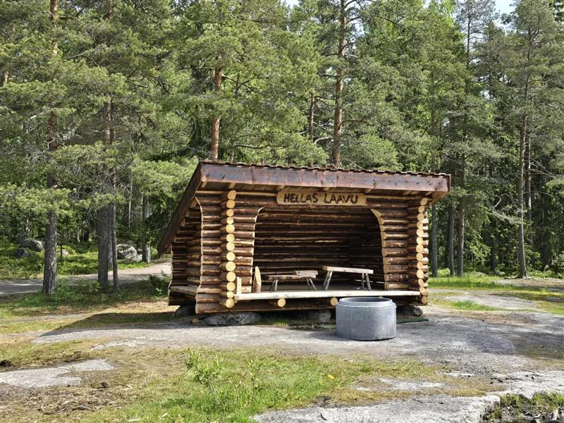 Hirsistä rakennettu laavu metsämaisemassa. Laavun sisällä kaksi puista penkkiä. Laavun yläosassa on kyltti, jossa lukee "Hellas Laavu".