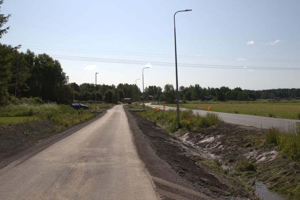 An asphalt-covered bicycle path runs through the countryside in bright daylight. On the left is forest, and on the right is farmland.
