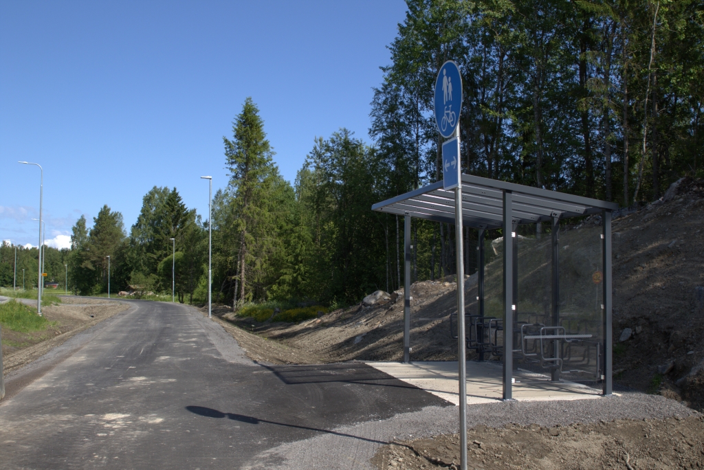 Pyöräpysäköintikatos metsän vieressä, asfaltoidun tien reunalla.