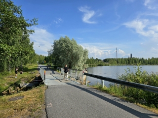 People walking on a bridge that crosses over water. In the background, there is a park and the sea.