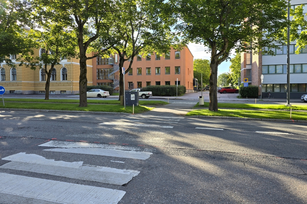 En övergång vid gatan i en grönskande omgivning, med ljusa byggnader i bakgrunden.