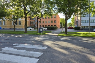 En övergång vid gatan i en grönskande omgivning, med ljusa byggnader i bakgrunden.