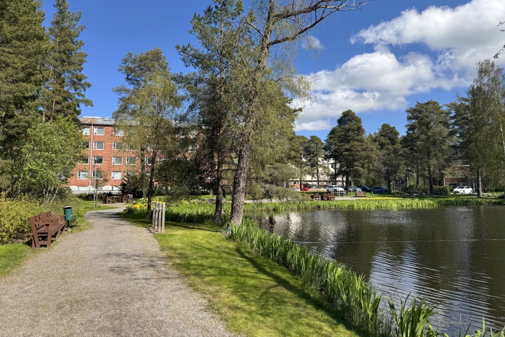 Puistomaisema, jossa kävelypolku kulkee lammen rantaa pitkin. Taustalla on punatiilinen rakennus.