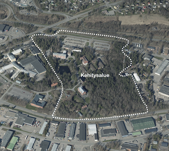Aerial view of a city area where a white dashed line marks a designated development area. The outlined area includes forest, several buildings, and parking areas. Surrounding the development area are industrial and commercial buildings as well as a street network.