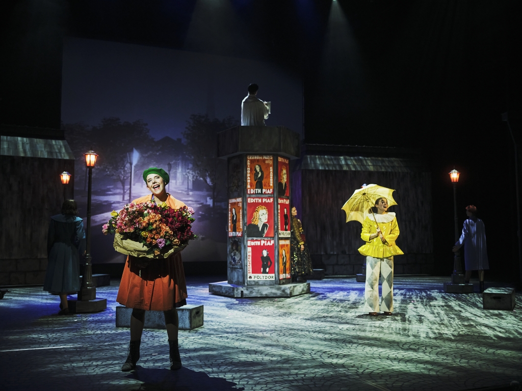 Kaksi henkilöä, toisella kukkakori kädessä ja toisella keltainen sateenvarjo ja sadetakki, esityskuva Piaf-musiikkinäytelmästä.