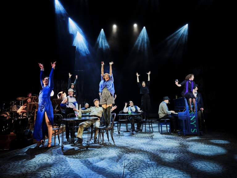 Kymmenkunta henkilöä roolihahmoissa näyttämöllä, osa istuu ja osa seisoo pöydällä kädet ylhäällä, oikealla piano, jonka päällä istuu henkilö, esityskuva Piaf-musiikkinäytelmästä.