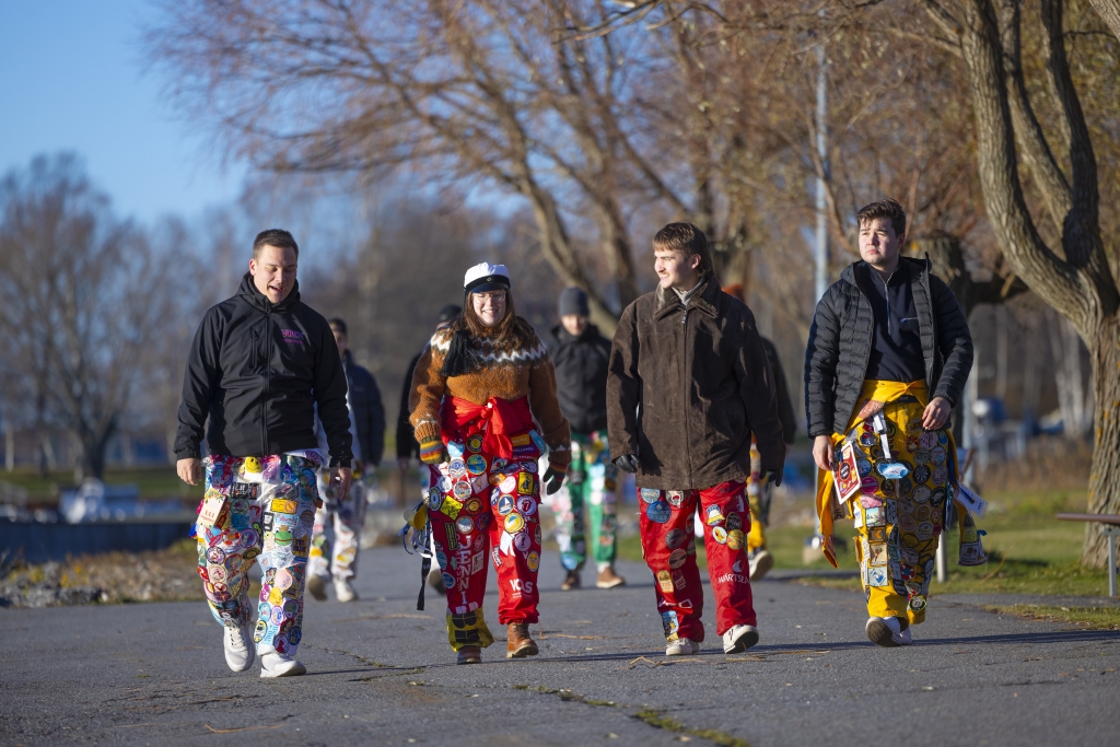 Four students walk outdoors on a sunny day wearing colorful student overalls covered with patches. Leafless trees can be seen in the background.