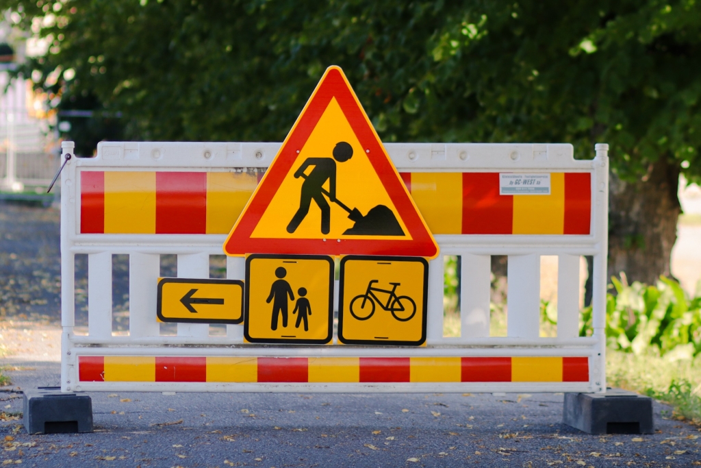 Construction site fence with a triangular roadwork sign, a pedestrian arrow sign, and a bicycle sign. In the background, there are trees.