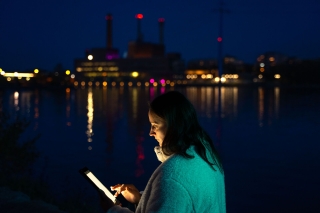Person using a tablet by the water at dusk, with illuminated industrial buildings and city lights reflecting on the surface.
