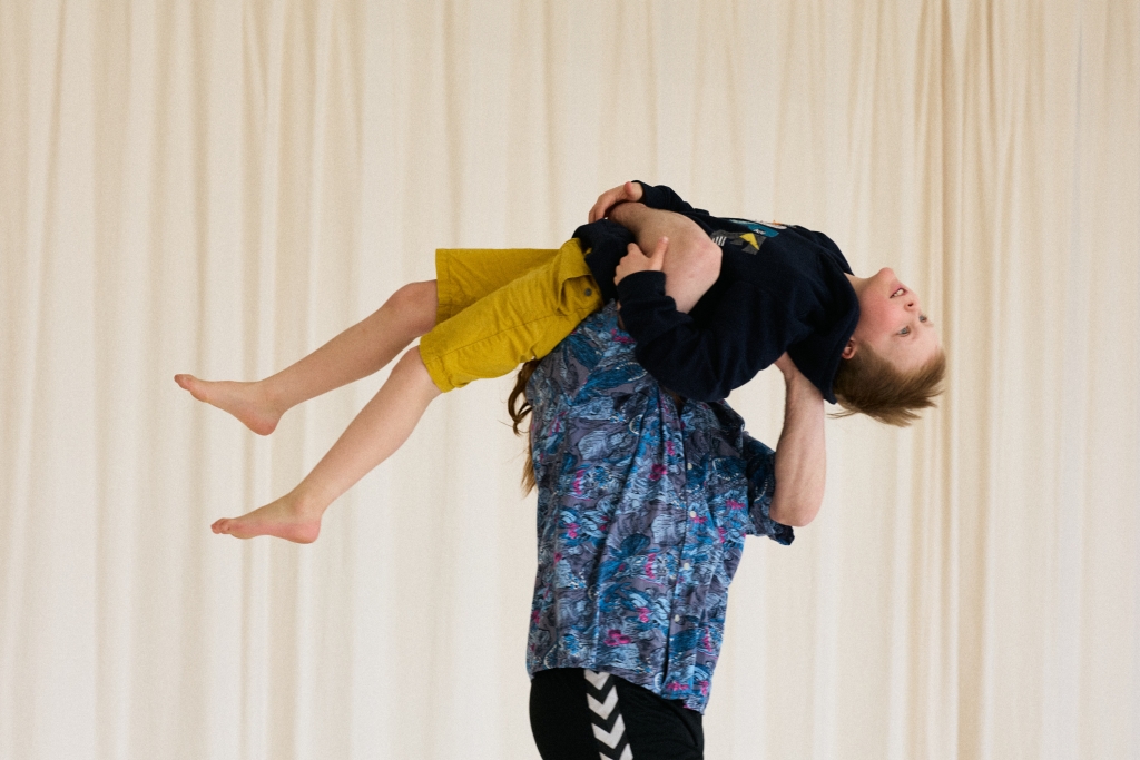 Kaksi henkilöä, aikuinen kantaa lasta harteillaan, takana vaaleat verhot.