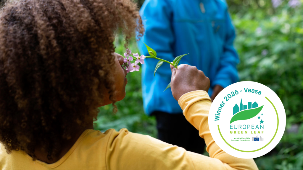 A person smells pink flowers in a green environment. The image also shows the logo of the European Green Leaf 2026 winner, Vaasa.