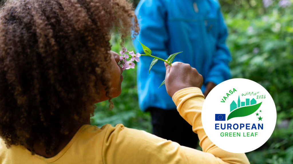 A person smells pink flowers in a green environment. The image also shows the logo of the European Green Leaf 2026 winner, Vaasa.