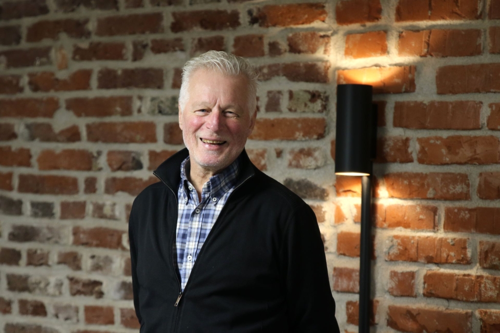 Hymyilevä ikääntynyt mies seisoo tiiliseinän edessä lämpimässä valossa.