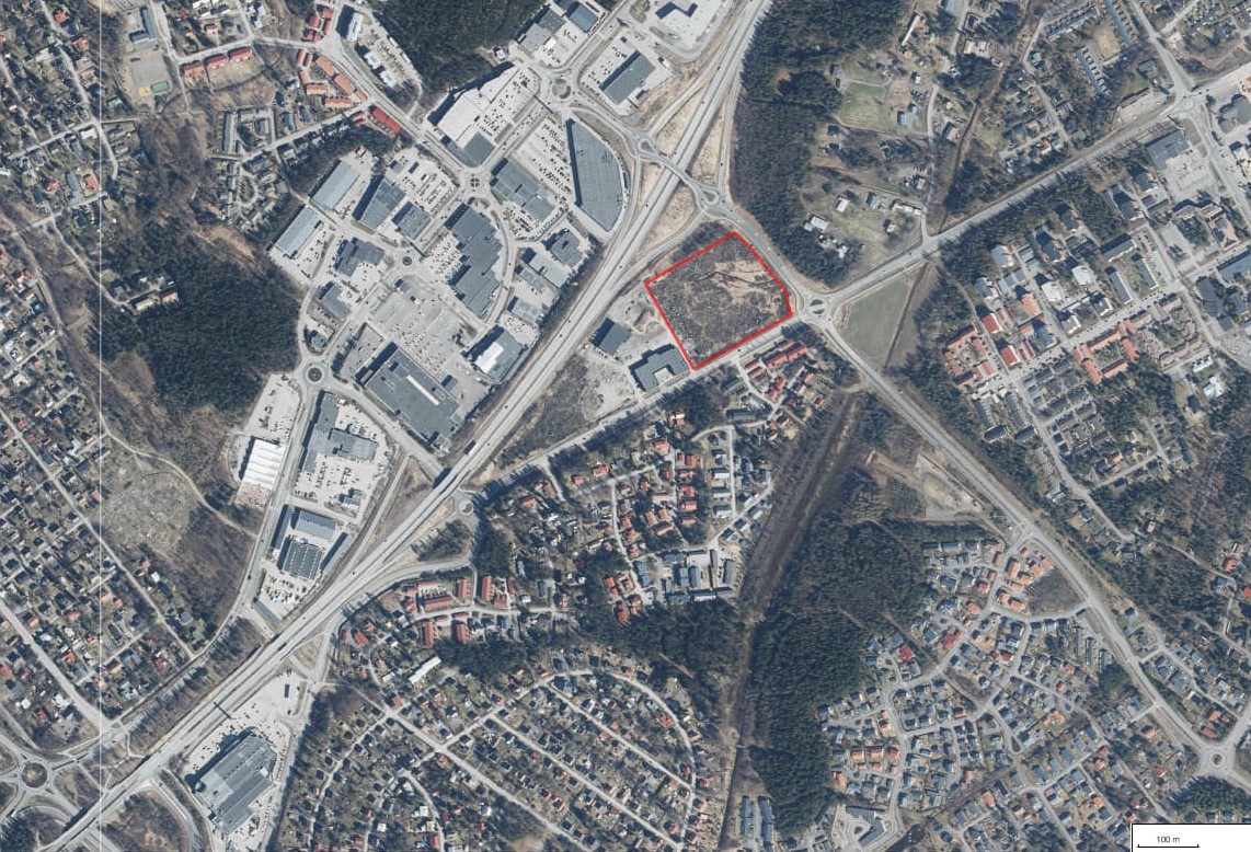 En tom tomt som ligger vid livligt trafikerade förbindelser mellan ett stort handelsområde och bostadsområden. Tjänster, köpcentrum och omfattande bostadsområden i närområdet omger tomten.