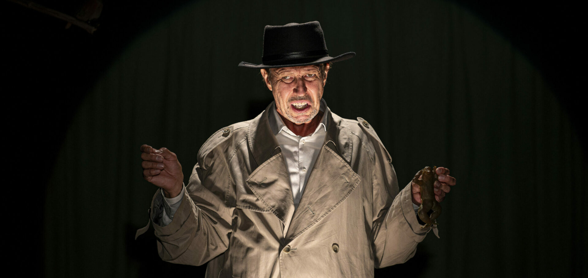 Näyttelijä katsoo kameraan, kädet sivuilla, päällä takki ja hattu.