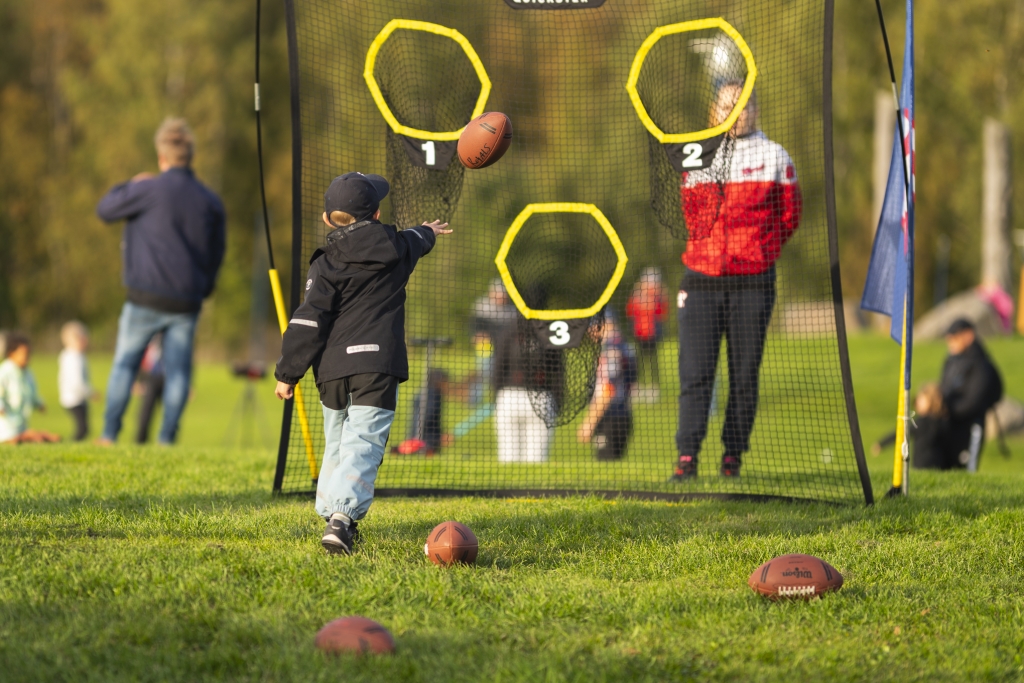 Lapsi heittää amerikkalaista jalkapalloa verkkoon, jossa on kolme merkittyä heittoaukkoa puistomaisessa ympäristössä.