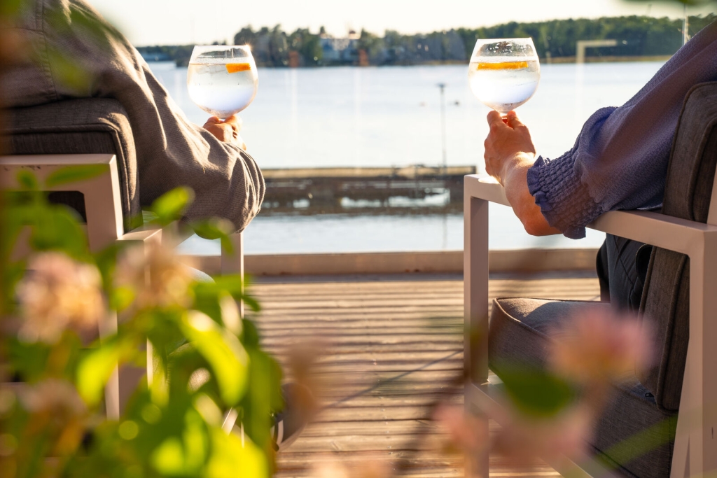 Kaksi ihmistä istuu terassilla ja pitää kädessään juomalaseja auringonpaisteessa, taustalla näkyy rauhallinen merimaisema.