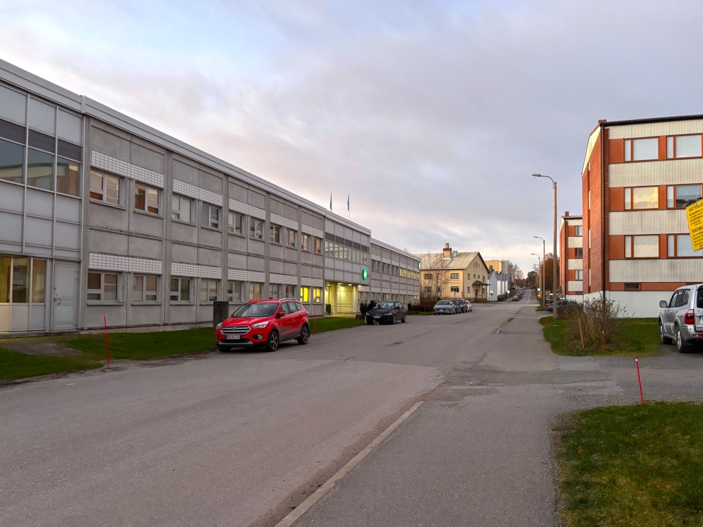 En gata med en kontorsbyggnad på ena sidan och ett hyreshus på den andra. Bilar står parkerade längs gatan.