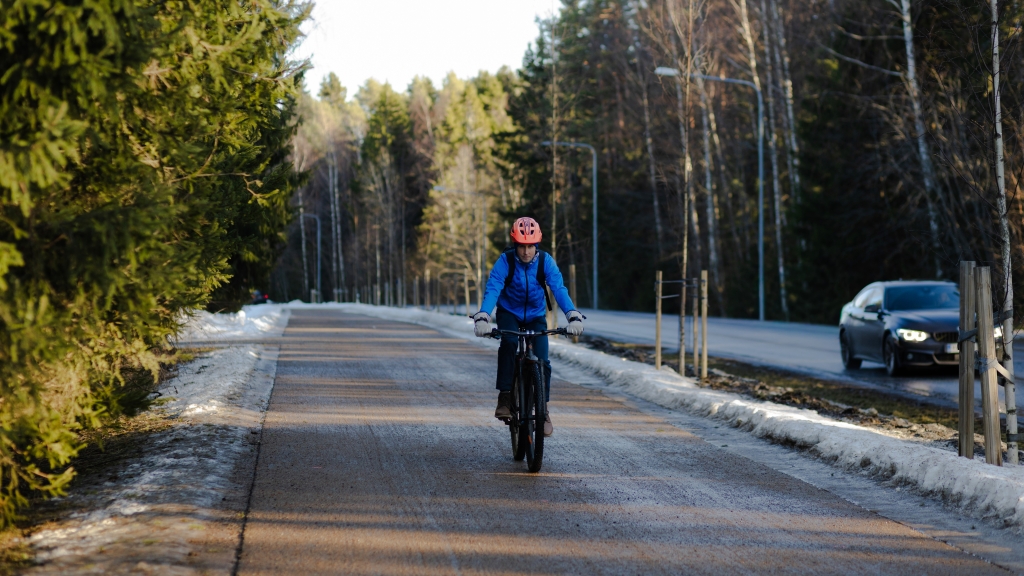 Henkilö pyöräilee keväisellä pyörätiellä metsän ja autotien vieressä.