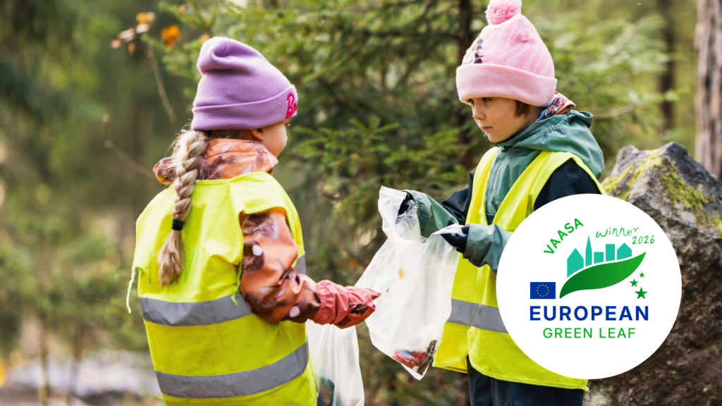 Kaksi lasta kerää roskia metsäisessä ympäristössä, päällään kirkkaat heijastinliivit ja lämpimät ulkovaatteet. Toinen pitää avointa keräyspussia, johon he ovat pudottamassa roskia.