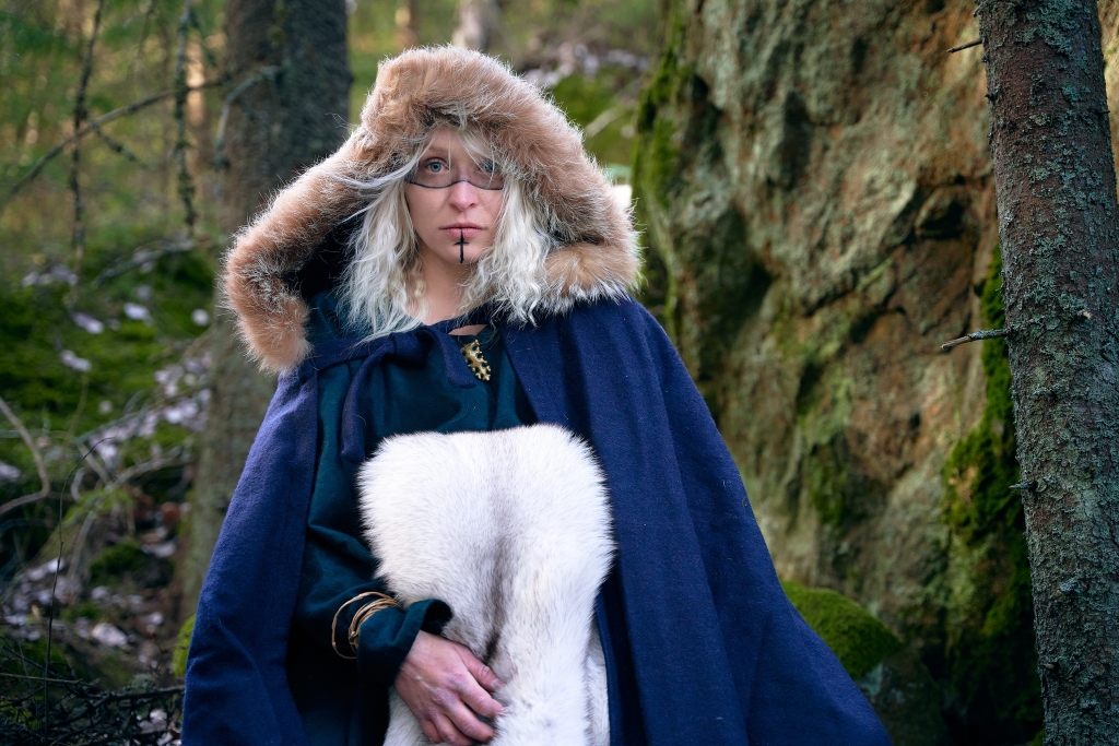 Kuvassa on viikingiksi pukeutunut nainen, jolla on sininen viitta ja huppu päässä. Hupussa on turkisreunus. Naisella on käsivarrella siniketusta tehty turkis ja kasvoille on maalattu mustia kuviointeja. Nainen katsoo kameraan.