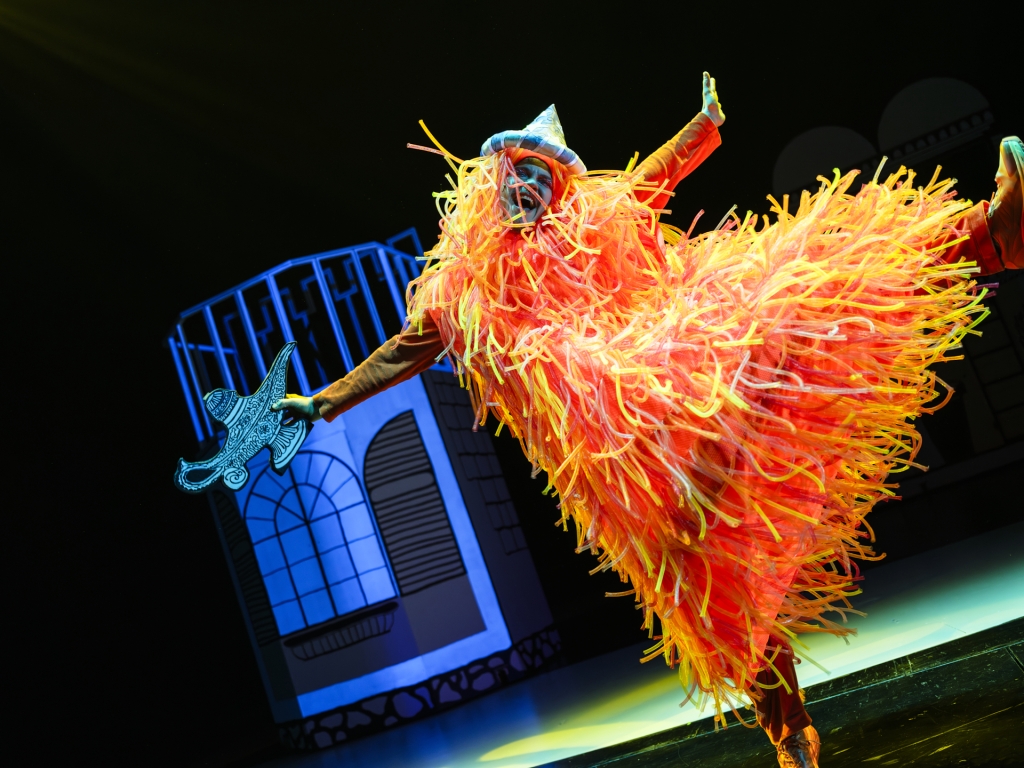 Henkilö oranssissa näyttävässä asussa tanssii näyttämöllä, esityskuva Aladdin ja taikalamppu -näytelmästä.