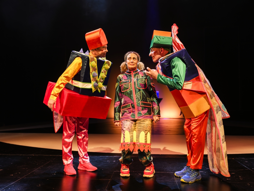 Kolme henkilöä näyttämöllä värikkäissä asuissa, kaksi reunimmaista kääntyneenä toisiinsa, keskellä henkilö huolestunut ilme kasvoillaan. Esityskuva Aladdin ja taikalamppu -näytelmästä.