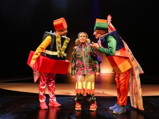 Kolme henkilöä näyttämöllä värikkäissä asuissa, kaksi reunimmaista kääntyneenä toisiinsa, keskellä henkilö huolestunut ilme kasvoillaan. Esityskuva Aladdin ja taikalamppu -näytelmästä.