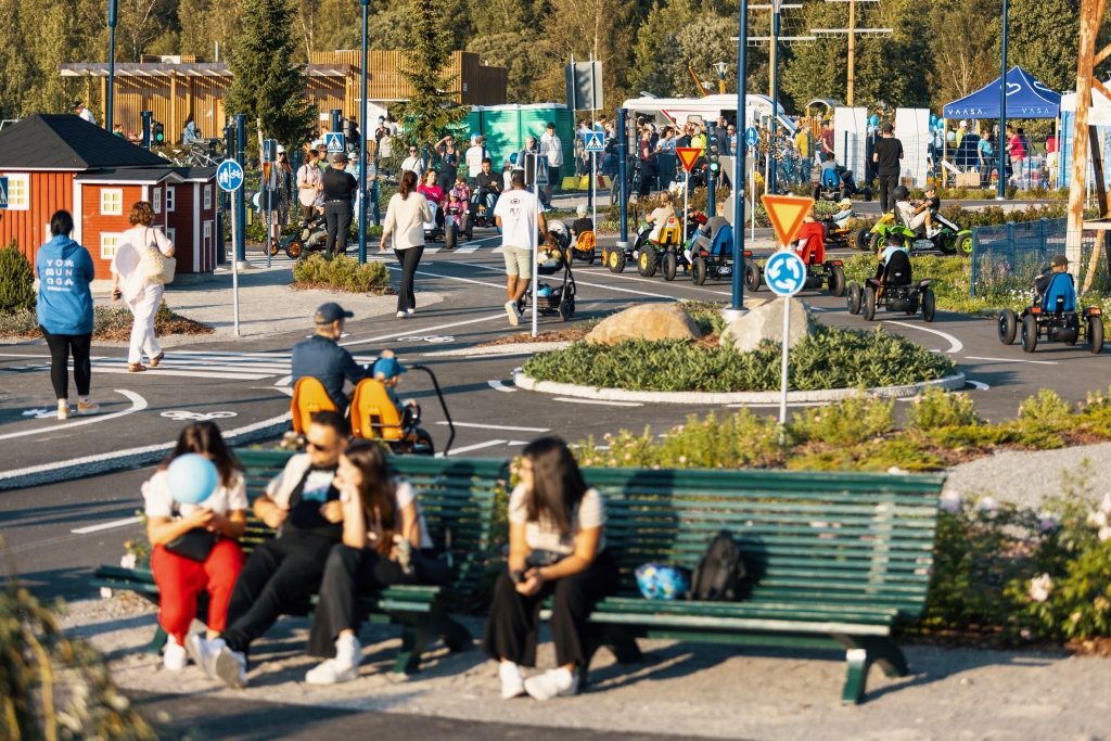 Puistoalue, jossa on paljon ihmisiä nauttimassa ulkoilmasta. Etualalla penkeillä istuu väkeä, ja taustalla lapsia leikkimässä polkuautoilla.