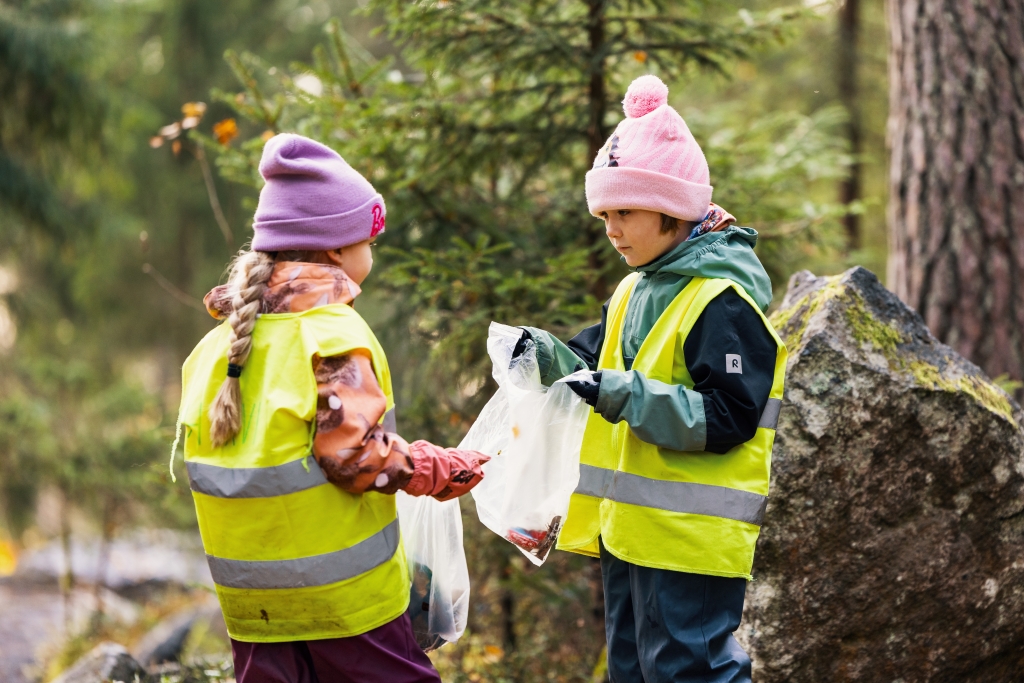 Två barn står i skogen och håller i genomskinliga plastpåsar fyllda med sopor.