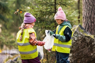 Två barn står i skogen och håller i genomskinliga plastpåsar fyllda med sopor.