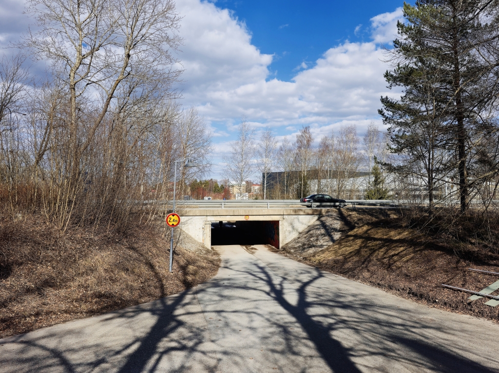 Vägen går under en låg bro. På bron finns en motorväg där man kör bil. Runt omkring finns träd och i bakgrunden syns en mulen himmel.