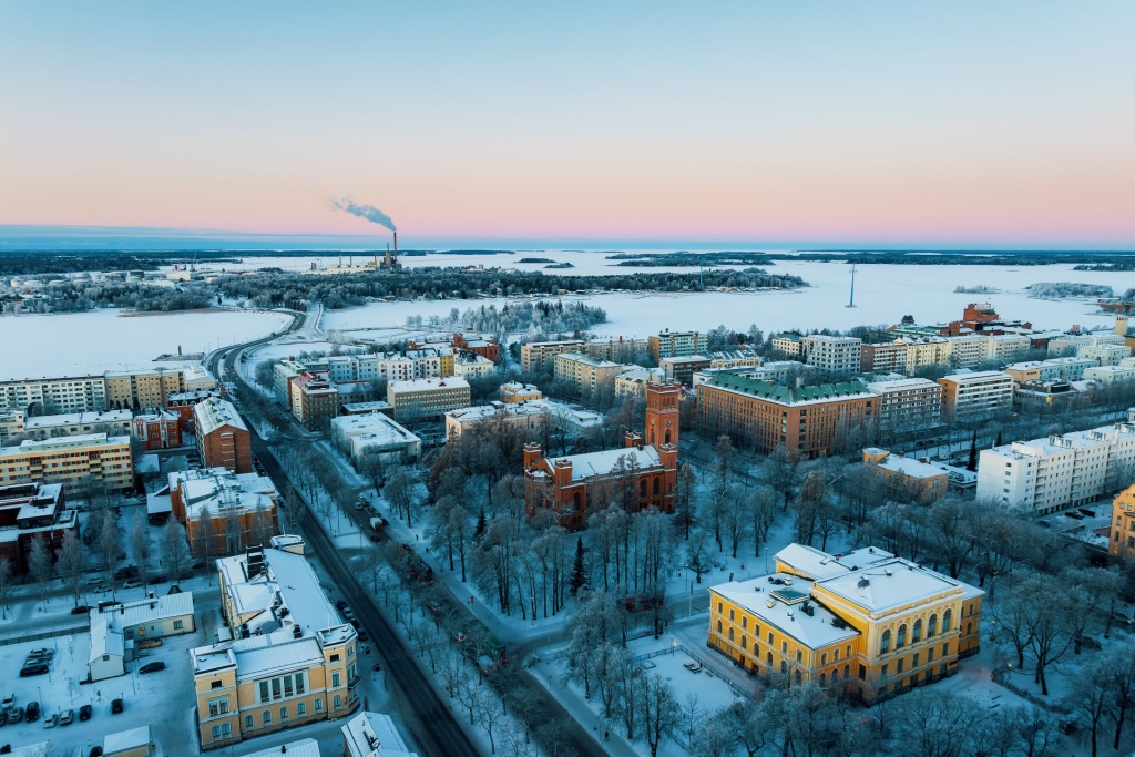 Ilmakuva talvisesta kaupunkimaisemasta meren rannalla: lumisia katuja ja rakennuksia, puistoalue sekä etäällä näkyvä teollisuuslaitos savupiippuineen auringonlaskun värjäämän taivaan alla.