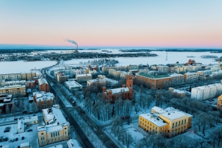Ilmakuva talvisesta kaupunkimaisemasta meren rannalla: lumisia katuja ja rakennuksia, puistoalue sekä etäällä näkyvä teollisuuslaitos savupiippuineen auringonlaskun värjäämän taivaan alla.