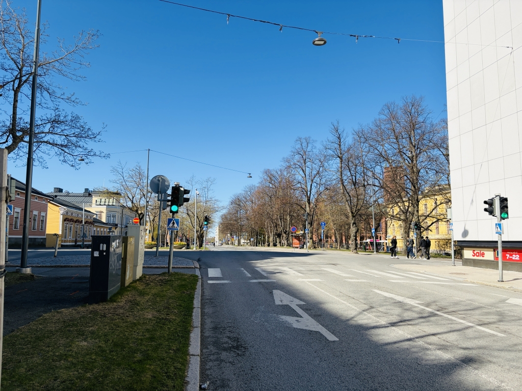 Leveä risteys ja suojatie, vihreät liikennevalot. Vaaleita rakennuksia molemmin puolin. Muutama jalankulkija ylittää tien oikealla.