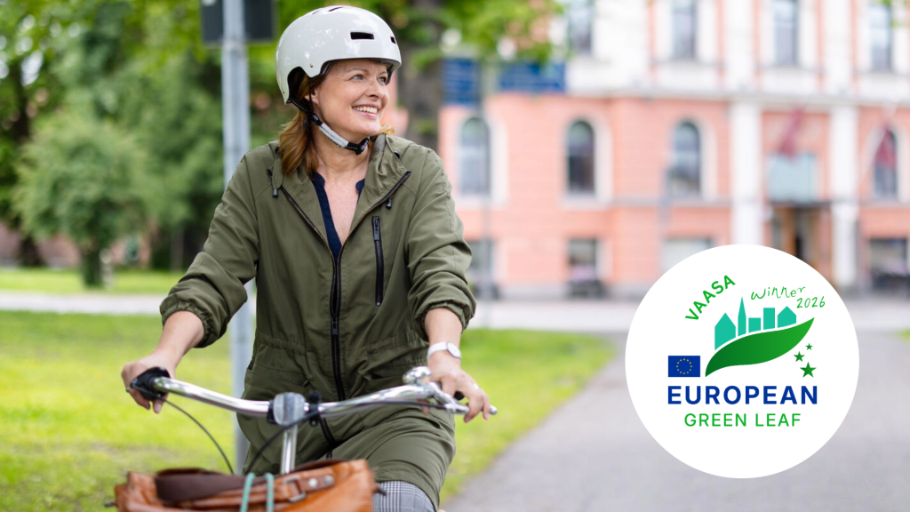A person riding a bicycle in an urban setting, wearing a helmet. Green park areas and residential buildings are visible in the background. In the lower right corner, the European Green Leaf logo appears with the text “Vaasa winner 2026”.