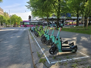 Rivi sähköpotkulautoja pysäköitynä kadun varteen. Taustalla linja-auto, vihreitä puita ja rakennuksia.