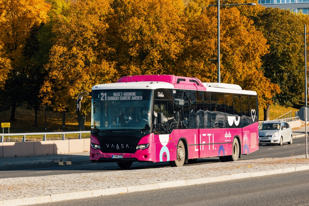 Vaaleanpunainen linja-auto ajaa maantiellä. Takana on lehtipuita ruskan väreissä.