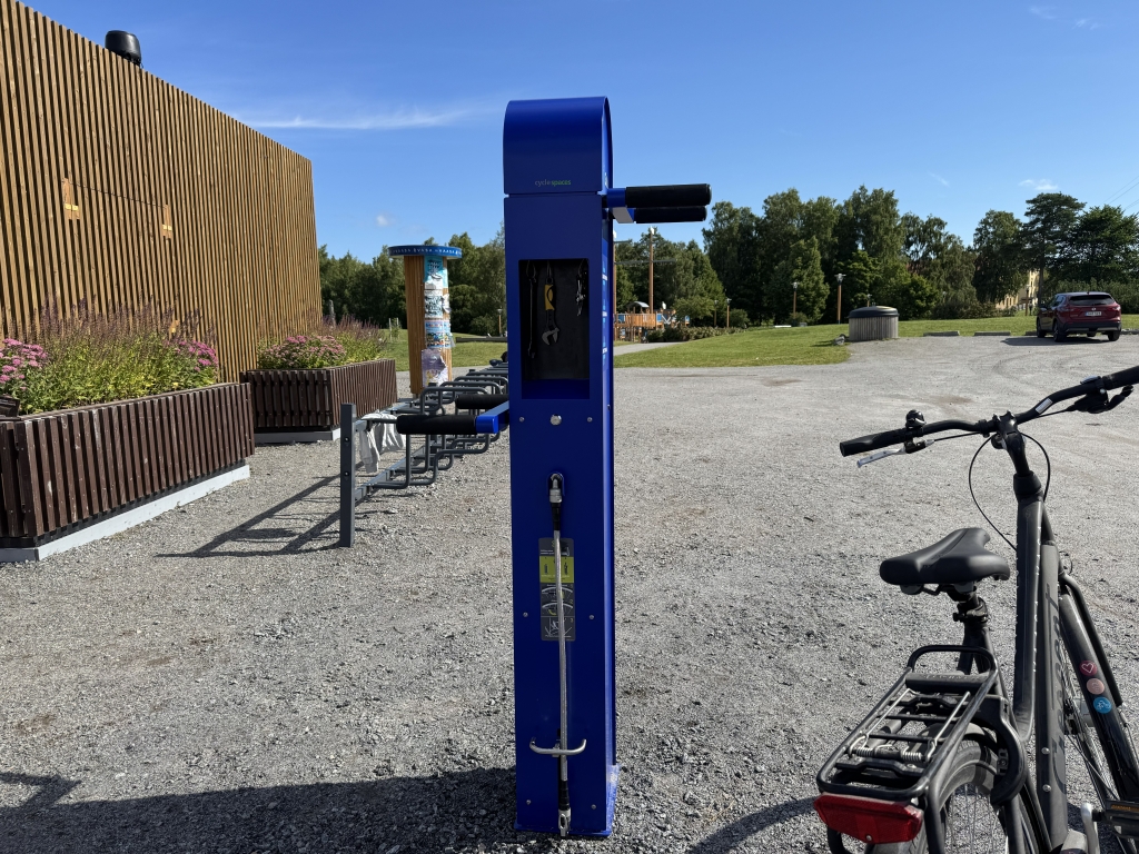 En blå cykelreparationställning bredvid kaféet i Metviken. Ställningen har en fördjupning med verktyg inuti samt stänger på båda sidor där man kan hänga upp cykeln. Under fördjupningen finns en cykelpump som drivs med en pedal.