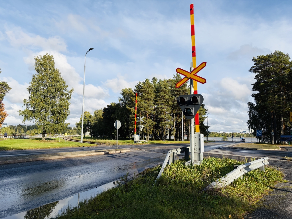 Rautatien tasoristeys, varoitusvalot ja puomit, taustalla puita ja pilvinen taivas.