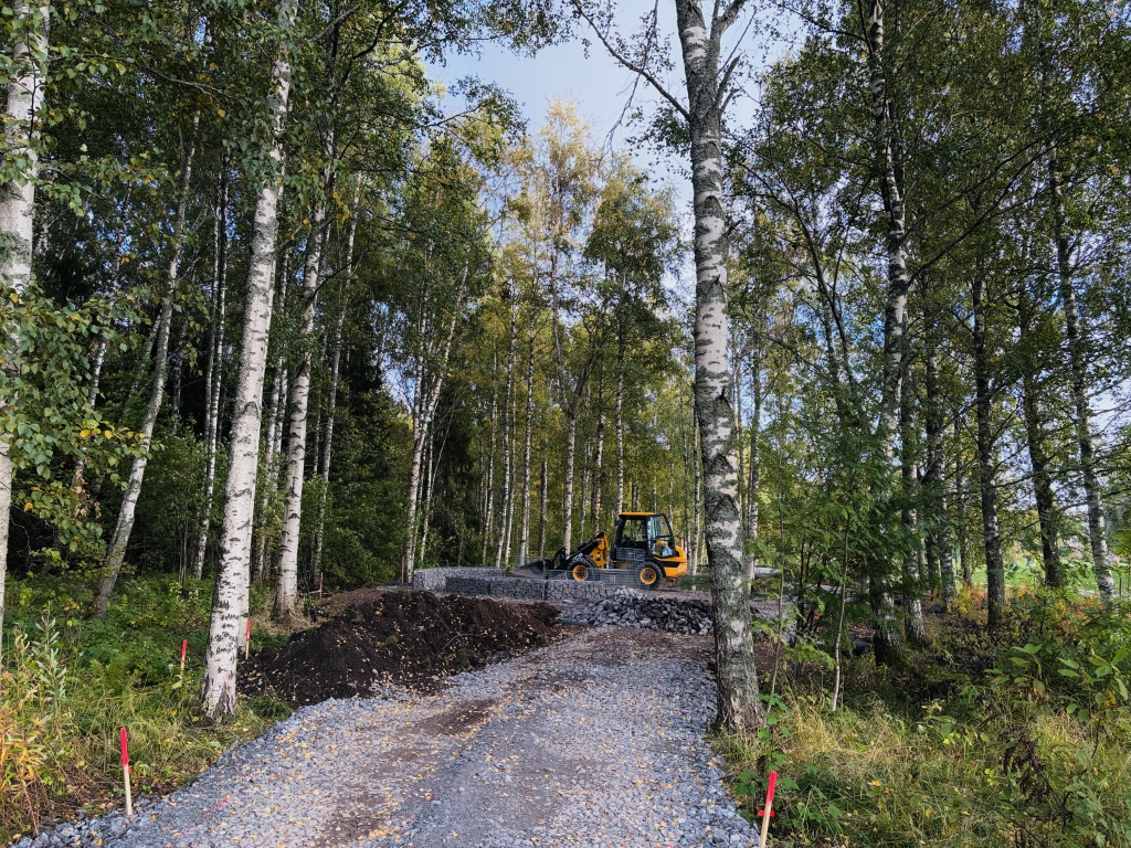 Koivumetsän keskellä kulkevalla sorapohjaisella reitillä tehdään rakennustöitä. Taustalla näkyy työkone ja rakenteilla oleva kivirakenne luonnonympäristössä.