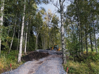 Koivumetsän keskellä kulkevalla sorapohjaisella reitillä tehdään rakennustöitä. Taustalla näkyy työkone ja rakenteilla oleva kivirakenne luonnonympäristössä.
