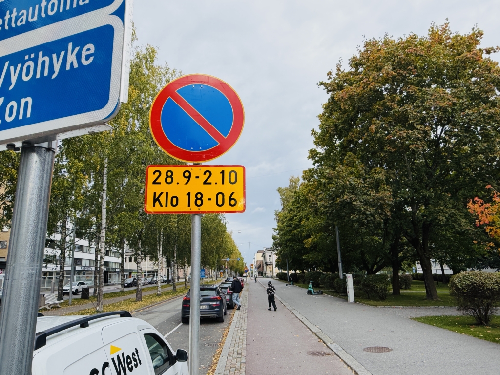 Pysäköintikieltomerkki kadun varressa. Merkin alla oleva lisäkilpi ilmoittaa pysäköintikiellon olevan voimassa 28.9.–2.10. kello 18.00–06.00. Taustalla näkyy kävely- ja pyörätie, puita, ajorata sekä pysäköityjä autoja.