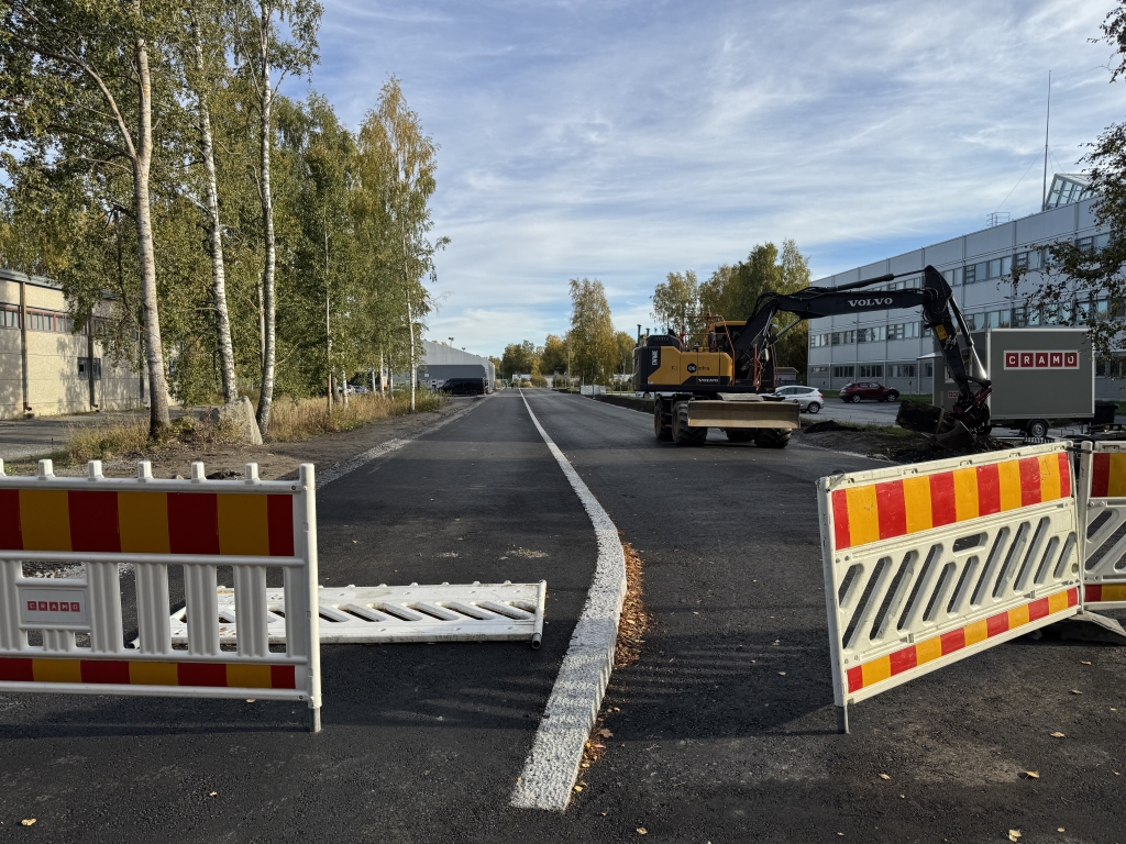 Asfaltoitu katu, jonka eteen on asetettu työmaa-aitoja. Kadun varrella näkyy kaivinkone, rakennuksia ja puita.