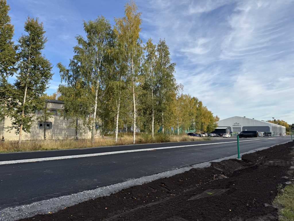Asfaltoitu katu, jonka toisella puolella kasvaa puita ja toisella puolella näkyy rakennuksia. Kadun reunoja viimeistellään.